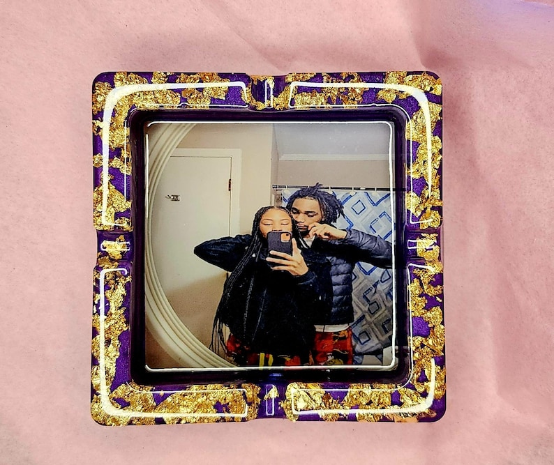 Op de afbeelding: Een paarse en gouden asbak met een foto van een stel erin. De foto toont een man en een vrouw die naar de camera lachen. De asbak heeft een witte rand en gouden vlokken overal.