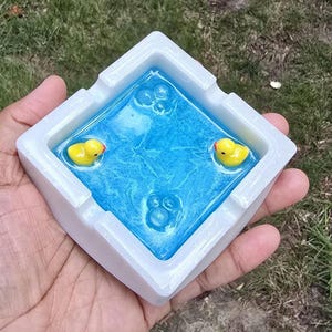 May include: A square ashtray with a white border and a blue interior. The blue interior has a water-like appearance with three yellow rubber duckies floating in it. The ashtray is held in a person's hand.