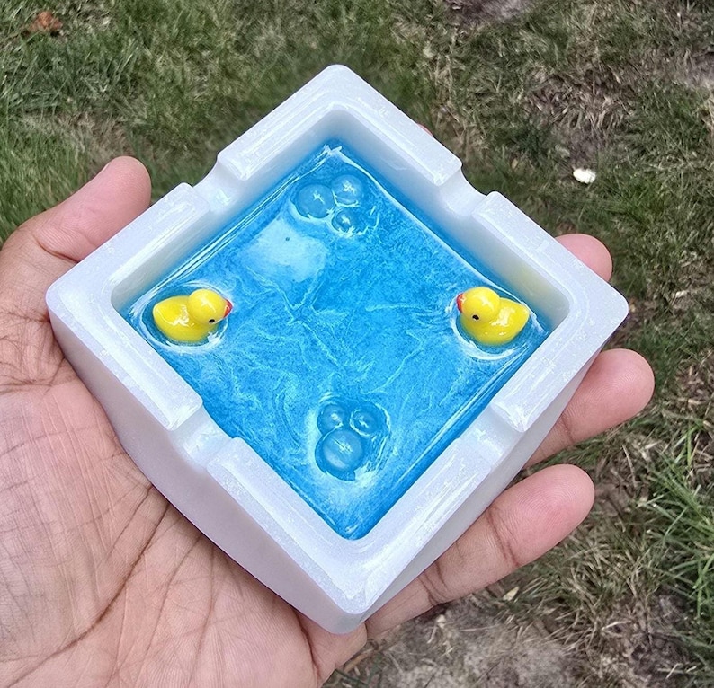 May include: A square ashtray with a white border and a blue interior. The blue interior has a water-like appearance with three yellow rubber duckies floating in it. The ashtray is held in a person's hand.