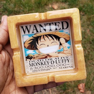 Op de afbeelding: Vierkante asbak met een gouden glitterafwerking. Het midden toont een "Wanted" poster van Monkey D. Luffy uit One Piece. De poster luidt "Dead or Alive" en vermeldt een premie van 300.000.000.