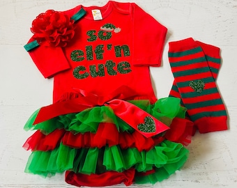 Puede incluir: Conjunto de bebé de Navidad rojo y verde con falda de tul, polainas y diadema. El body dice "So elf'n cute" en purpurina verde.
