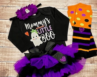 Może przedstawiać: Czarno-fioletowy strój dla niemowlęcia na Halloween z długim body z napisem "Mommy's Little Boo" i grafiką ducha, czarną tiulową spódniczką z fioletowymi falbankami oraz czarno-pomarańczowymi, paskowanymi getrami w kropki.