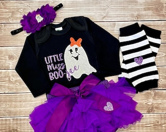 Może przedstawiać: Czarny, długorękawny śpioszek z błyszczącym białym duchem i napisem "Little Miss Boo-jee". Śpioszek jest zestawiony z fioletową tiulową spódniczką z fioletową satynową kokardką, fioletową opaską na głowę z białym duchem i czarno-białymi, prążkowanymi getrami z fioletowym sercem.