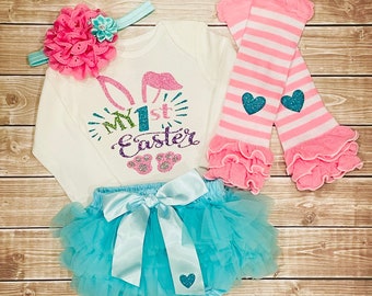 May include: A baby girl's Easter outfit with a white long-sleeved shirt that says "My 1st Easter" with glitter bunny ears, a turquoise tulle tutu with a light blue satin bow, and pink and white striped leg warmers with a ruffle trim.