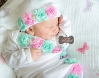 May include: A newborn baby wearing a white gown with a pink and mint green flower trim. The baby is sleeping in a wicker basket lined with a white blanket with pink butterfly designs. The baby is wearing a white hat with a pink and mint green flower trim.