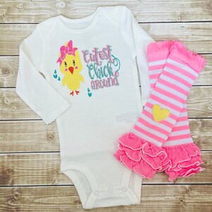 May include: White long-sleeved bodysuit with a yellow chick graphic and the text "Cutest Chick Around". The bodysuit is paired with pink and white striped leg warmers with a yellow heart.