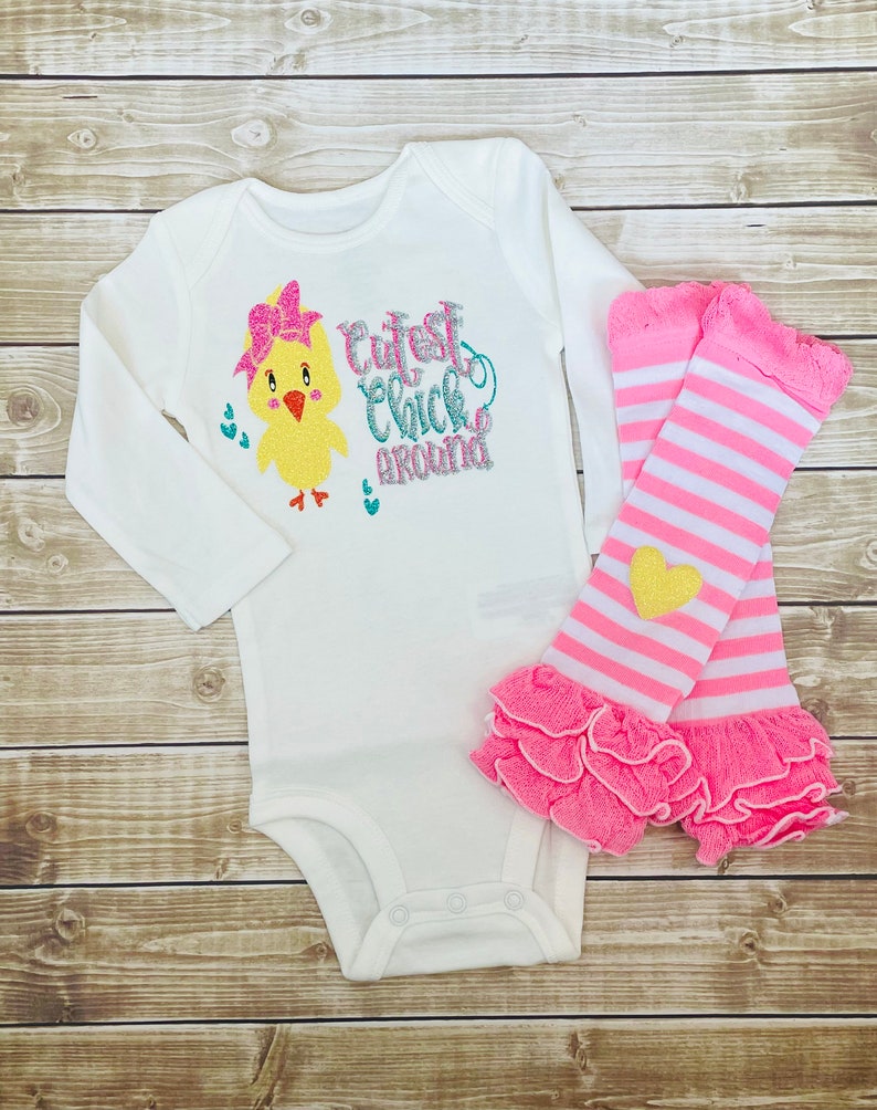 May include: White long-sleeved bodysuit with a yellow chick graphic and the text "Cutest Chick Around". The bodysuit is paired with pink and white striped leg warmers with a yellow heart.