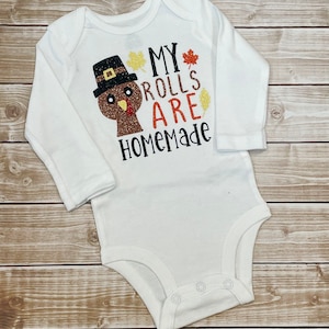May include: White long-sleeved baby bodysuit featuring a Thanksgiving turkey design and the words "MY ROLLS ARE HOMEMADE". The turkey has a brown body, black hat, and orange beak. The text is in black, orange, and gold glitter.