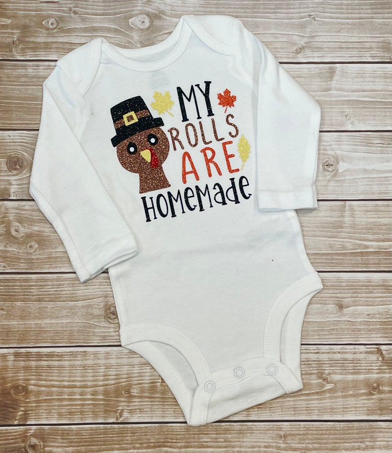 May include: White long-sleeved baby bodysuit featuring a Thanksgiving turkey design and the words "MY ROLLS ARE HOMEMADE". The turkey has a brown body, black hat, and orange beak. The text is in black, orange, and gold glitter.