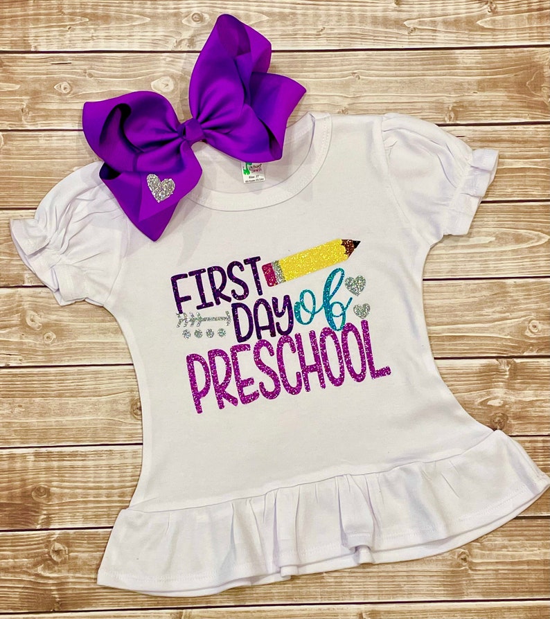 May include: White t-shirt with a purple, yellow, and blue glitter design that says "First Day of Preschool" with a yellow pencil graphic. A purple bow with a silver glitter heart is on the shirt.