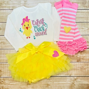 May include: A yellow tutu with a yellow satin bow and a pink heart with glitter. A white long-sleeved onesie with a yellow chick with a pink bow and the text "Cutest Chick Around". Pink and white striped leg warmers with a pink ruffle.