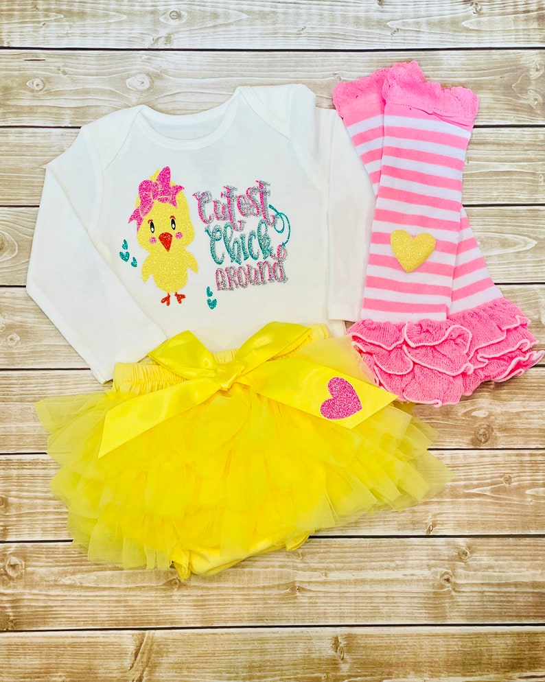 May include: A yellow tutu with a yellow satin bow and a pink heart with glitter. A white long-sleeved onesie with a yellow chick with a pink bow and the text "Cutest Chick Around". Pink and white striped leg warmers with a pink ruffle.