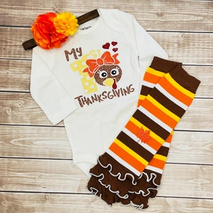 May include: White long-sleeve onesie with the text "My 1st Thanksgiving" and a glittery turkey graphic. The onesie is paired with brown and orange striped leggings with a brown ruffle at the bottom.
