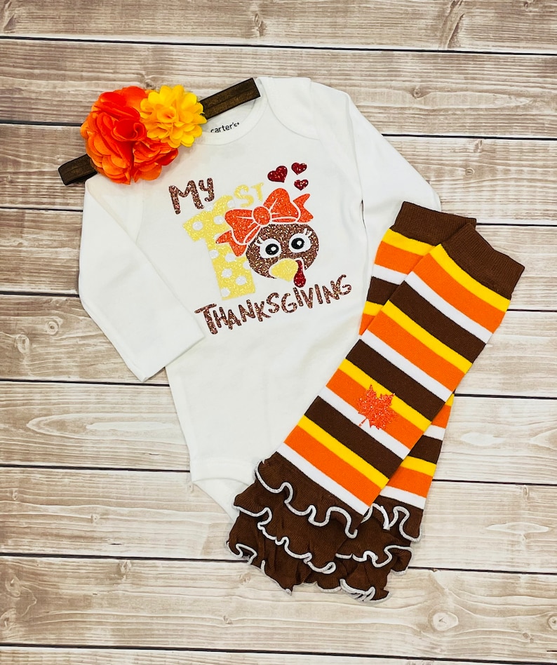 May include: White long-sleeve onesie with the text "My 1st Thanksgiving" and a glittery turkey graphic. The onesie is paired with brown and orange striped leggings with a brown ruffle at the bottom.