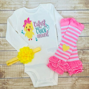May include: A white long-sleeved onesie with a yellow chick wearing a pink bow and the text "Cutest Chick Around". The onesie is paired with pink and white striped leg warmers with a yellow heart and a yellow flower headband.