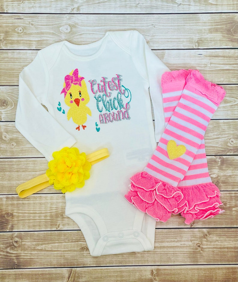May include: A white long-sleeved onesie with a yellow chick wearing a pink bow and the text "Cutest Chick Around". The onesie is paired with pink and white striped leg warmers with a yellow heart and a yellow flower headband.