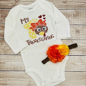 May include: White long-sleeved onesie with the text "My 1st Thanksgiving" and a glittery turkey with a bow. A brown headband with an orange and yellow flower is included.