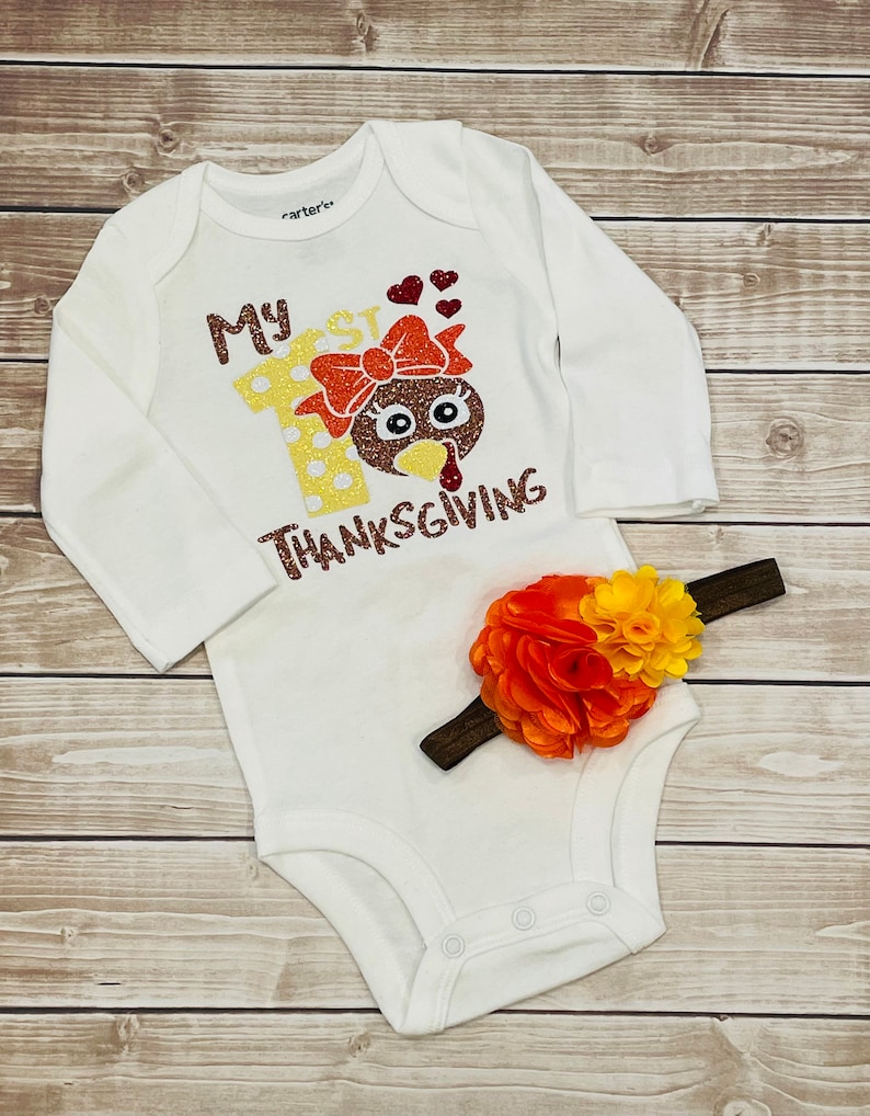 May include: White long-sleeved onesie with the text "My 1st Thanksgiving" and a glittery turkey with a bow. A brown headband with an orange and yellow flower is included.