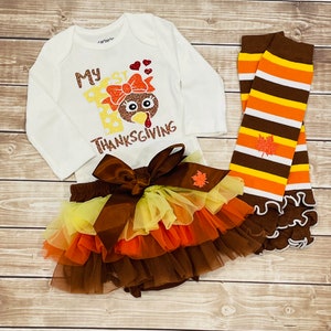 May include: A white onesie with the text "My 1st Thanksgiving" and a glitter turkey graphic. A brown tulle tutu with yellow, orange, and brown layers. A pair of brown, orange, and white striped leg warmers with a glitter leaf.