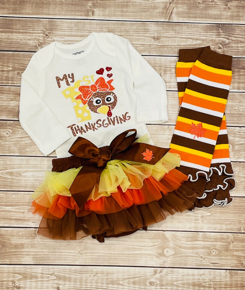 May include: A white onesie with the text "My 1st Thanksgiving" and a glitter turkey graphic. A brown tulle tutu with yellow, orange, and brown layers. A pair of brown, orange, and white striped leg warmers with a glitter leaf.