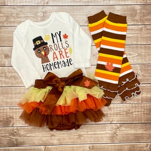 May include: A white baby bodysuit with a turkey graphic and the text "My Rolls Are Homemade." The set includes a layered tutu skirt with a large bow, and striped leg warmers with leaf accents. The outfit is in autumnal colours.
