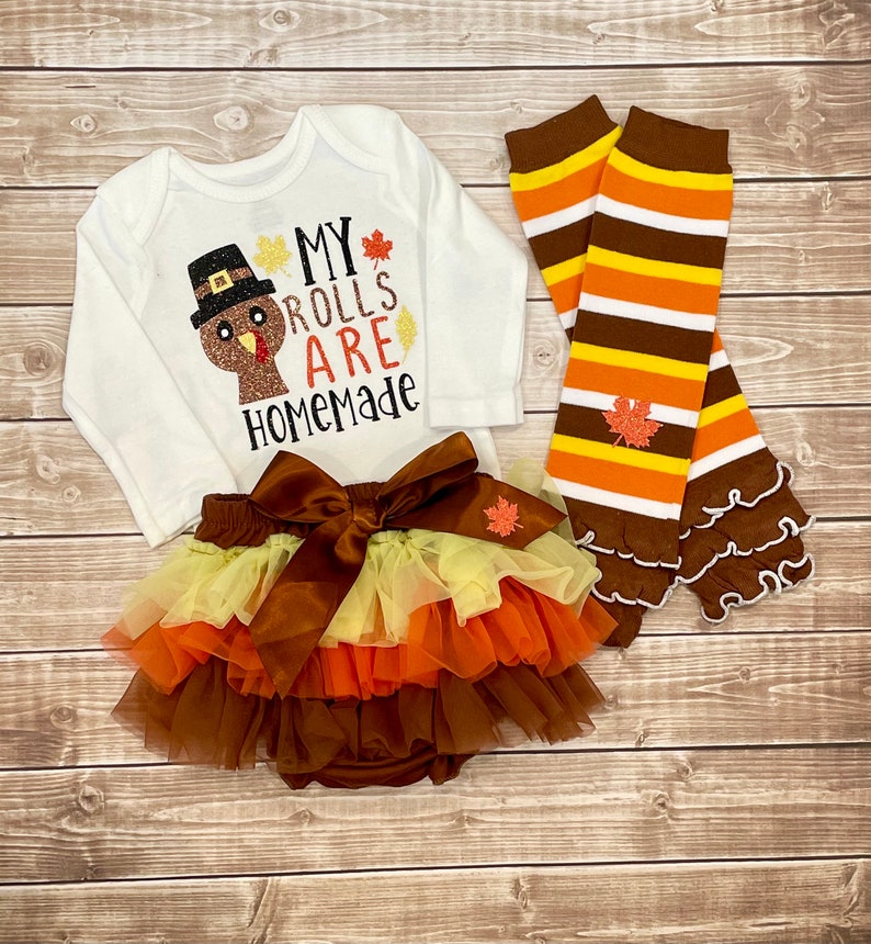 May include: A white baby bodysuit with a turkey graphic and the text "My Rolls Are Homemade." The set includes a layered tutu skirt with a large bow, and striped leg warmers with leaf accents. The outfit is in autumnal colours.