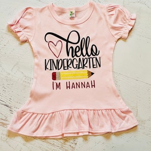 May include: Pink ruffle-bottom t-shirt with a black and white graphic that says "hello kindergarten" with a red heart and a yellow pencil with the text "I'm Hannah".