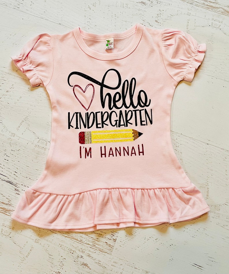 May include: Pink ruffle-bottom t-shirt with a black and white graphic that says "hello kindergarten" with a red heart and a yellow pencil with the text "I'm Hannah".