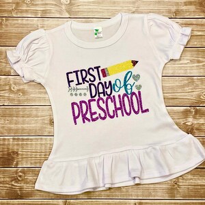 May include: White t-shirt with a ruffled hem, featuring a glittery design that says "First Day of Preschool" with a yellow pencil and hearts.