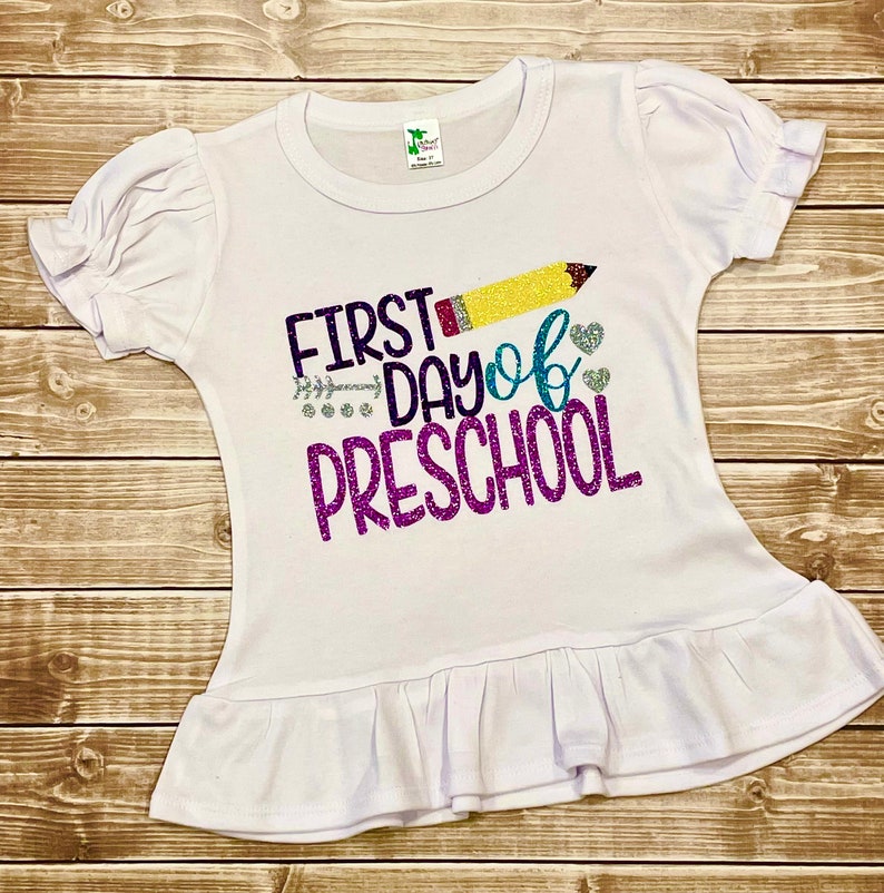 May include: White t-shirt with a ruffled hem, featuring a glittery design that says "First Day of Preschool" with a yellow pencil and hearts.