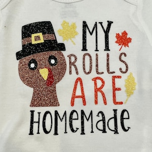 May include: White baby bodysuit featuring a glittery turkey design with a black pilgrim hat. The text "MY ROLLS ARE HOMEMADE" is printed in black and red, with gold and red leaf accents.