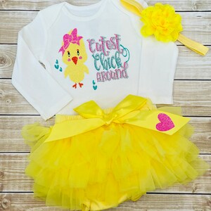 May include: A yellow tutu with a white bodysuit featuring a yellow chick with a pink bow and the text "Cutest Chick Around". The outfit includes a yellow headband with a yellow flower.