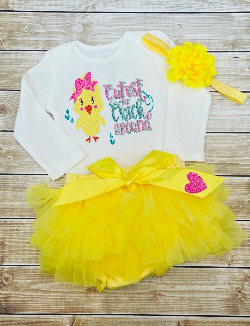 May include: A yellow tutu with a white bodysuit featuring a yellow chick with a pink bow and the text "Cutest Chick Around". The outfit includes a yellow headband with a yellow flower.
