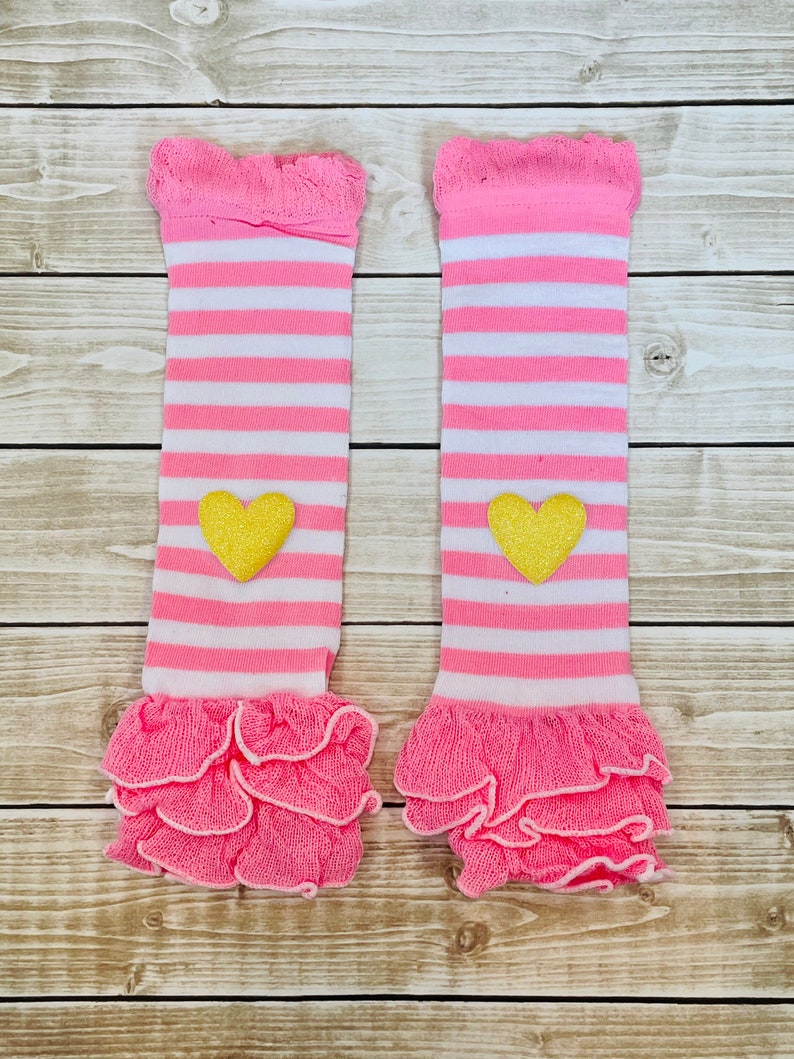 May include: A pair of pink and white striped leg warmers with a yellow heart on each. The leg warmers have a ruffled edge.