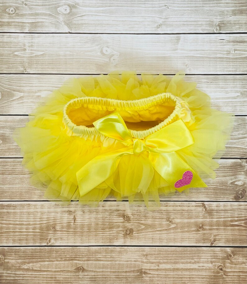 May include: A yellow tulle tutu with a yellow satin bow and a pink glitter heart.
