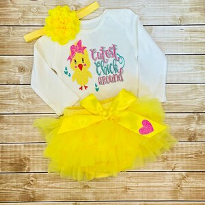 May include: A yellow baby outfit with a long-sleeved shirt featuring a yellow chick with a pink bow and the text "Cutest Chick Around". The outfit also includes a yellow tulle tutu with a pink heart and a yellow headband with a yellow flower.