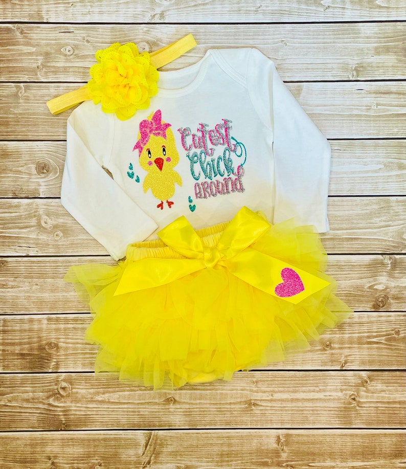 May include: A yellow baby outfit with a long-sleeved shirt featuring a yellow chick with a pink bow and the text "Cutest Chick Around". The outfit also includes a yellow tulle tutu with a pink heart and a yellow headband with a yellow flower.