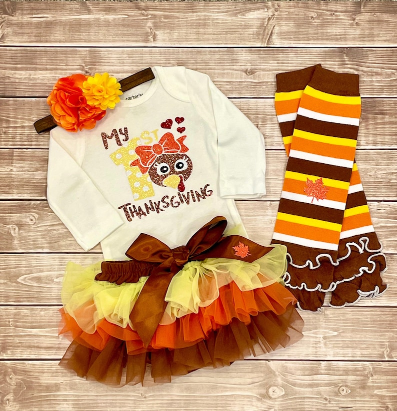May include: A white long-sleeved onesie with the text "My 1st Thanksgiving" and a glittery turkey graphic. The onesie is paired with a brown, orange, and yellow tulle tutu and brown and orange striped leg warmers with a ruffle trim.