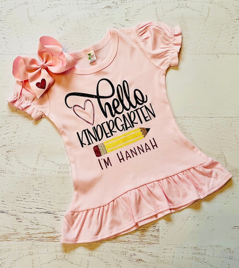 May include: Pink t-shirt with a black and glitter design that says "Hello Kindergarten" with a pencil and the text "I'm Hannah".