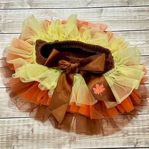 May include: A layered tutu skirt in autumnal colours, featuring shades of brown, orange, and yellow. The skirt has a brown elastic waistband with a large bow and a small leaf applique.