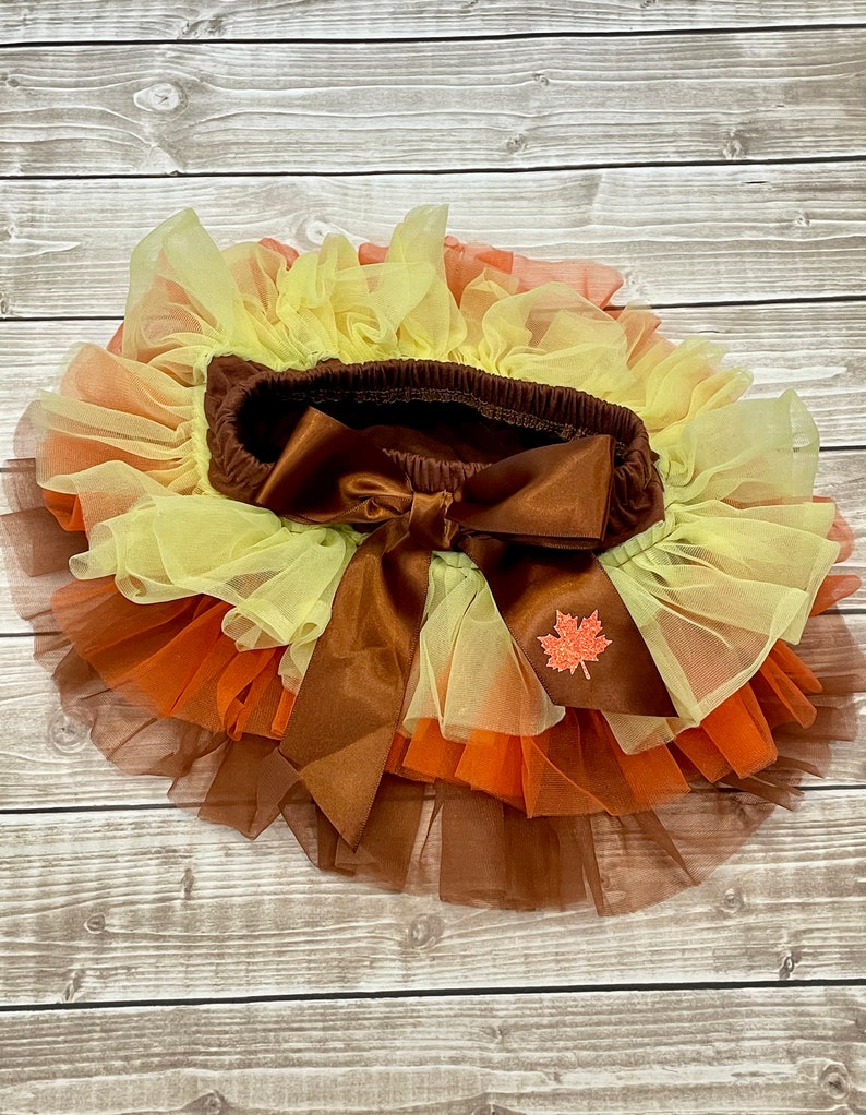 May include: A layered tutu skirt in autumnal colours, featuring shades of brown, orange, and yellow. The skirt has a brown elastic waistband with a large bow and a small leaf applique.