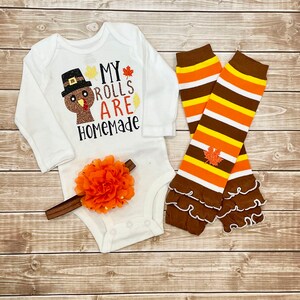 May include: A white baby bodysuit with long sleeves, featuring a turkey design and the words "MY ROLLS ARE HOMEMADE." The set includes a brown and orange floral headband and striped leg warmers with frills.
