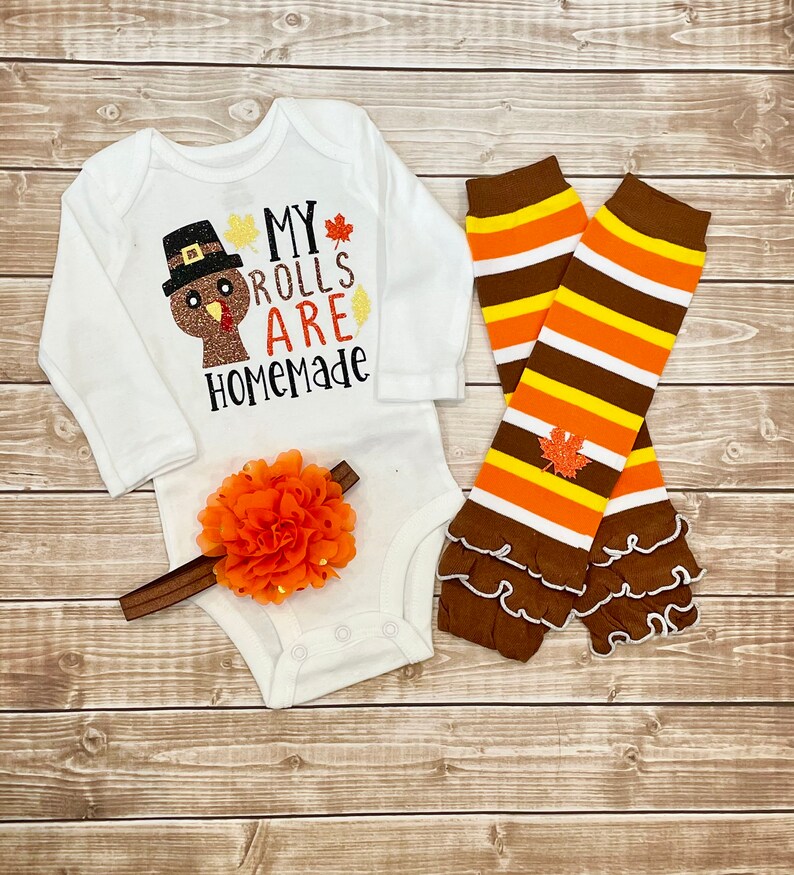 May include: A white baby bodysuit with long sleeves, featuring a turkey design and the words "MY ROLLS ARE HOMEMADE." The set includes a brown and orange floral headband and striped leg warmers with frills.