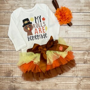 May include: A white baby bodysuit with long sleeves, featuring a turkey design and the words "My Rolls Are Homemade." The set includes a layered tulle skirt in brown, orange, and yellow, plus a floral headband.