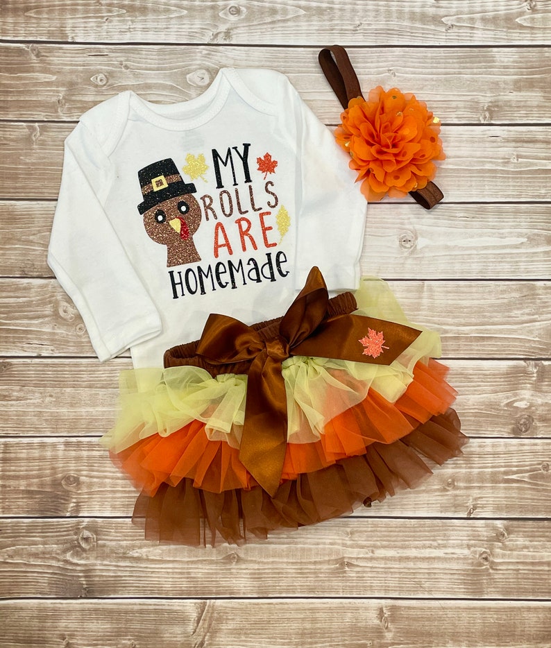 May include: A white baby bodysuit with long sleeves, featuring a turkey design and the words "My Rolls Are Homemade." The set includes a layered tulle skirt in brown, orange, and yellow, plus a floral headband.