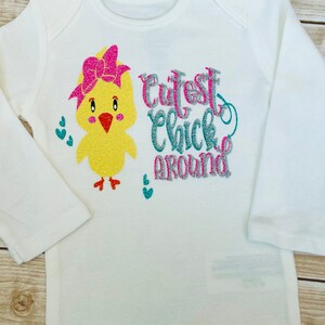 May include: White long-sleeved onesie with a yellow chick wearing a pink bow and the text "Cutest Chick Around" in pink, silver, and teal glitter.