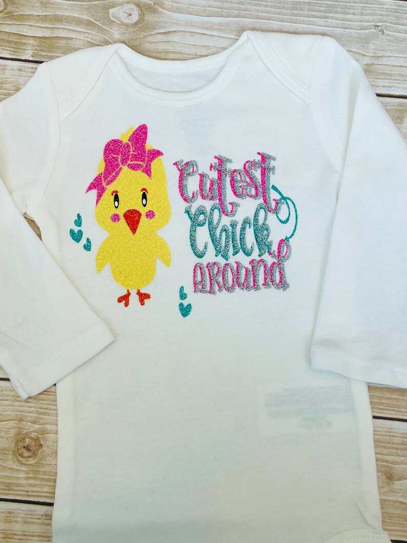 May include: White long-sleeved onesie with a yellow chick wearing a pink bow and the text "Cutest Chick Around" in pink, silver, and teal glitter.