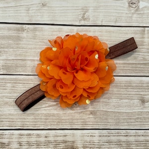 May include: A vibrant orange flower headband with gold polka dots. The flower is made of layered fabric petals and is attached to a brown elastic band. Suitable for babies and young children.