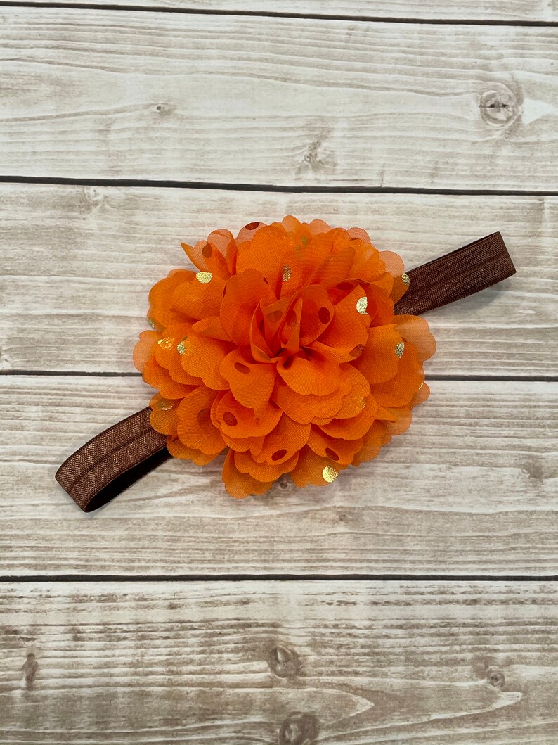 May include: A vibrant orange flower headband with gold polka dots. The flower is made of layered fabric petals and is attached to a brown elastic band. Suitable for babies and young children.