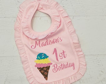 May include: Pink ruffled bib with the text "Madison's 1st Birthday" and a glitter ice cream cone graphic.